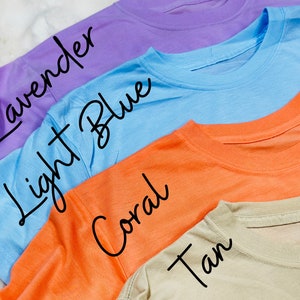 May include: Four colorful t-shirts in lavender, light blue, coral, and tan are displayed. The names of the colors are written in cursive on each shirt. The shirts are made of a soft material and have a crew neck.