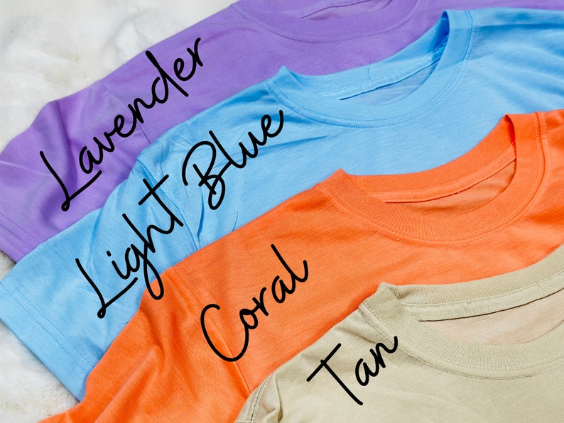 May include: Four colorful t-shirts in lavender, light blue, coral, and tan are displayed. The names of the colors are written in cursive on each shirt. The shirts are made of a soft material and have a crew neck.