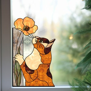 Op de afbeelding: Een raamdecoratie van gekleurd glas met een kat die aan een feloranje klaproos ruikt. De kat is bruin, oranje en wit met zwarte vlekken. De bloem heeft een groene stengel en bladeren.