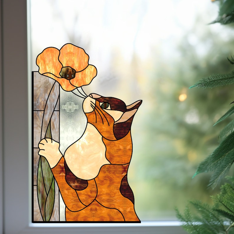 Op de afbeelding: Een raamdecoratie van gekleurd glas met een kat die aan een feloranje klaproos ruikt. De kat is bruin, oranje en wit met zwarte vlekken. De bloem heeft een groene stengel en bladeren.