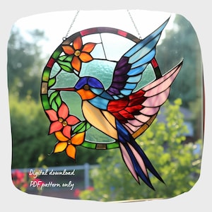 May include: A stained glass hummingbird with colorful wings and a flower design. The hummingbird is in a circular frame with a chain hanging from the top. The background is a blurry image of green foliage.