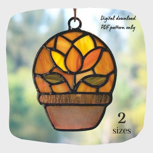 Op de afbeelding: Gebrandschilderde glazen suncatcher in de vorm van een bloem in een pot, met tinten oranje, geel en groen. De pot heeft een bruine basis en een metalen lus om op te hangen. Tekst: "Digital download PDF pattern only" en "2 sizes".