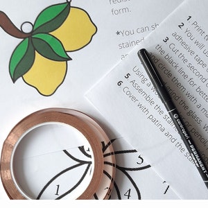 May include: A close-up of a craft project with instructions for making a stained glass lemon design. The instructions include using copper tape, a black marker, and a clear adhesive tape. The image also shows a roll of copper tape and a black marker.