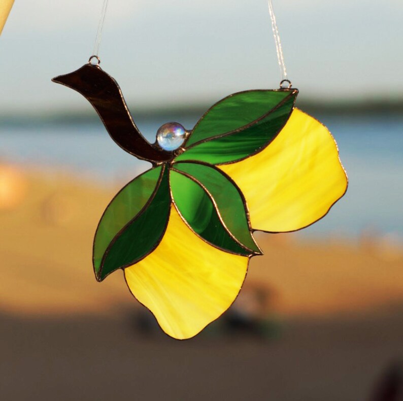 May include: A stained glass suncatcher featuring two yellow lemons with green leaves and a brown bird. The suncatcher is hanging from a clear string.