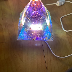May include: A pyramid-shaped lamp with a clear resin base filled with gold flakes and a blue, green, and purple gradient. The lamp is lit from within, casting a warm glow.