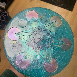 May include: A round, teal-colored decorative piece with a geometric design. The piece features a central Metatron's Cube pattern, surrounded by circles and geometric shapes. The design is accented with iridescent and glitter effects.