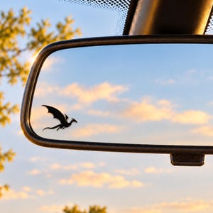May include: A black dragon silhouette decal applied to the lower left corner of a rectangular vehicle rearview mirror. The mirror reflects a bright blue sky with soft, golden-hued clouds during sunset, showing the dragon in flight.