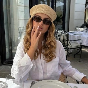 Peut inclure: Une personne portant un béret en paille beige et des lunettes de soleil sombres, assise à une table de restaurant. Elle porte une chemise blanche boutonnée. Une assiette blanche avec un motif de feuille verte et un verre à vin sont sur la table.