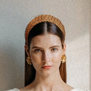 Könnte beinhalten: Eine Frau mit einem braunen gehäkelten Stirnband und goldenen, blumenförmigen Ohrringen.