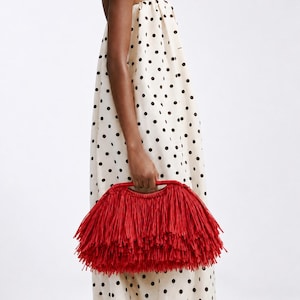 May include: A strapless cream-coloured dress featuring a black polka dot pattern and a loose, flowing silhouette. The garment is paired with a vibrant red handbag covered in long, thin, layered fringe strands and a structured top handle.