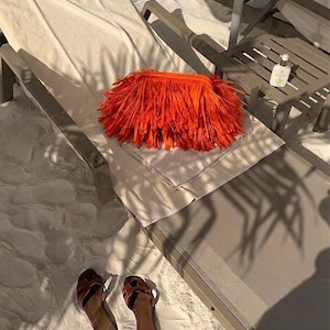 Könnte beinhalten: Eine orangefarbene Clutch-Tasche mit Fransen liegt auf einer Sonnenliege mit einem weißen Handtuch. Braune Ledersandalen liegen auf dem Sand. Eine Flasche Lotion steht auf einem Beistelltisch. Die Szene ist von Sonnenlicht durchflutet, was auf einen entspannten Tag am Strand hindeutet.