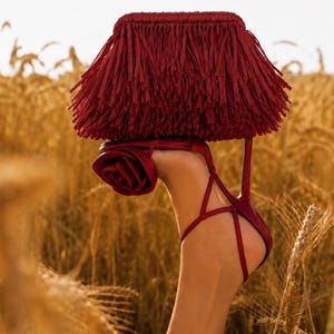 May include: A deep red clutch bag covered in long fringe tassels paired with a matching deep red high-heeled sandal featuring a prominent fabric rose detail on the side and thin ankle straps that wrap around the heel and foot area.