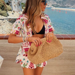 May include: A woman wearing a white and pink floral patterned shirt with a black bikini top and a straw bag.