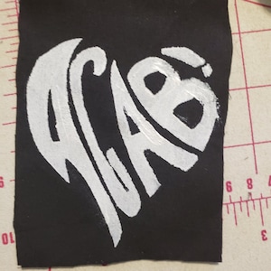 Peut inclure: Un patch en tissu noir avec un motif en forme de cœur blanc. Le cœur contient les lettres "ACAB" dans une police stylisée.