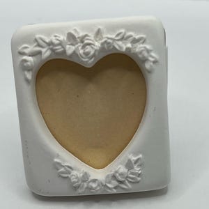 May include: A white, heart-shaped picture frame with floral embellishments. The frame has a rectangular shape with a heart cutout in the center. The floral details are arranged around the heart and on the corners.