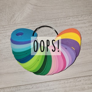 May include: A colourful fan of paper circles in various shades of blue, green, pink, purple, and orange. The circles are attached to a metal ring. The text "OOPS!" is printed on a white rectangle in front of the fan.