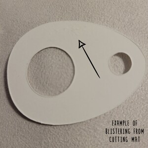 May include: A white, egg-shaped cutting mat with two circular cutouts and an arrow pointing to a small imperfection. The text "EXAMPLE OF BLISTERING FROM CUTTING MAT" is visible at the bottom of the image.