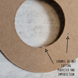 May include: Close-up of a tan cardboard cutout with a circular hole. The text "EXAMPLE OF NOT CUTTING PERFECTLY AND IMPERFECTION" is written in black. Arrows point to imperfections in the cut, highlighting the handmade nature.