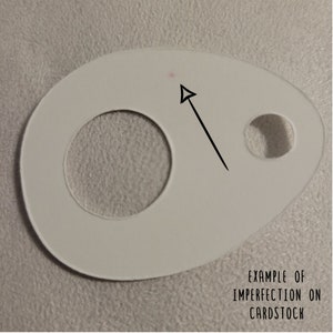 May include: A white cardstock shape with a large circular hole and a smaller hole. An arrow points to a small pink dot, with the text "EXAMPLE OF IMPERFECTION ON CARDSTOCK" below.