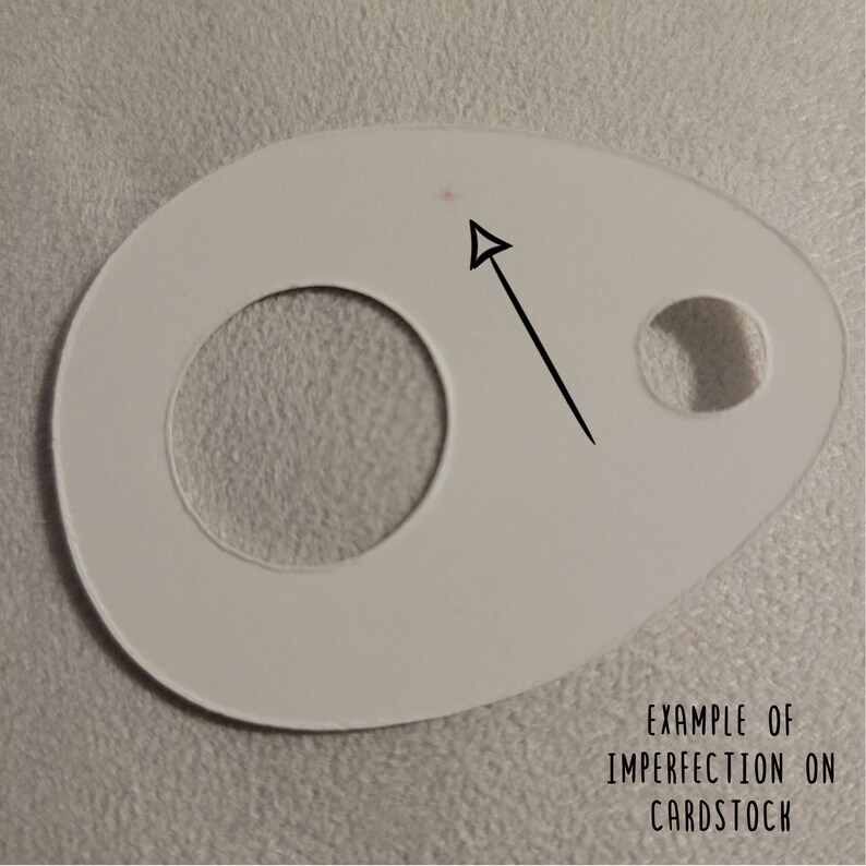 May include: A white cardstock shape with a large circular hole and a smaller hole. An arrow points to a small pink dot, with the text "EXAMPLE OF IMPERFECTION ON CARDSTOCK" below.