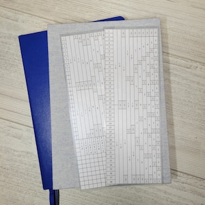 Pode incluir: Duas folhas de plástico brancas com grades numeradas, uma sobre a outra. As folhas estão em um caderno azul com fundo branco.