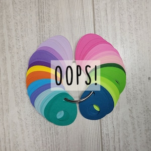 May include: A colour wheel with a variety of colours, including purple, pink, green, blue, and orange. The colour wheel is attached to a metal ring. The text "OOPS!" is printed on a white label.