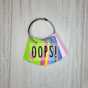 May include: A collection of colorful cards attached to a silver metal ring. The front card is lime green with the word "OOPS!" in black. Other cards are pink, blue, yellow, and white.