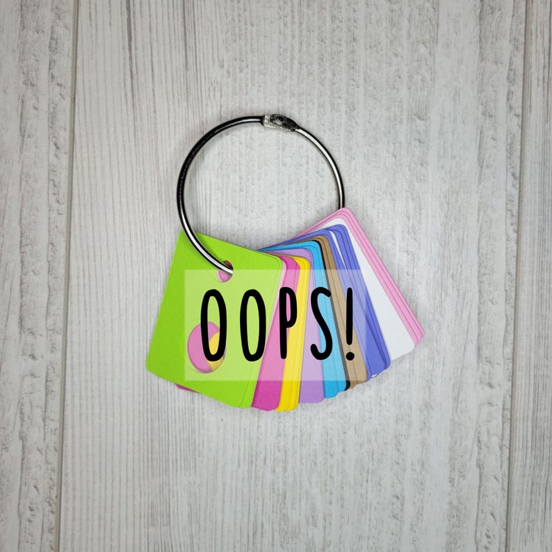 May include: A collection of colorful cards attached to a silver metal ring. The front card is lime green with the word "OOPS!" in black. Other cards are pink, blue, yellow, and white.