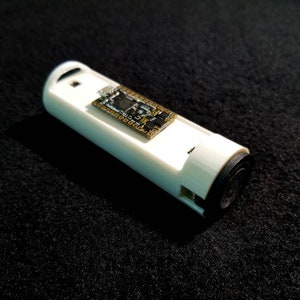 May include: A white, cylindrical electronic device with a small circuit board attached. The device has a black end cap and a small hole at the top. The device is resting on a black, textured surface.