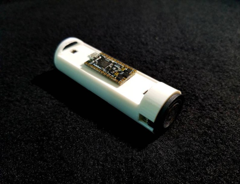 May include: A white, cylindrical electronic device with a small circuit board attached. The device has a black end cap and a small hole at the top. The device is resting on a black, textured surface.