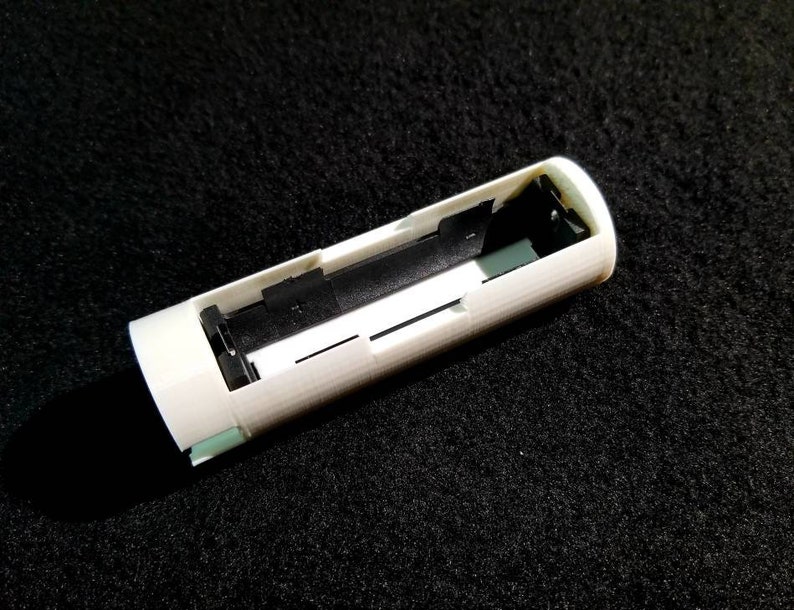 May include: A white, cylindrical object with a rectangular opening, revealing a black interior and small green accents. The object appears to be a component or housing for an electronic device, possibly a battery holder.