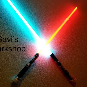 May include: Two illuminated lightsabers, one blue and one red, are crossed against a textured wall. The text "Savi's Workshop" is displayed on the wall. The lightsabers have black handles.
