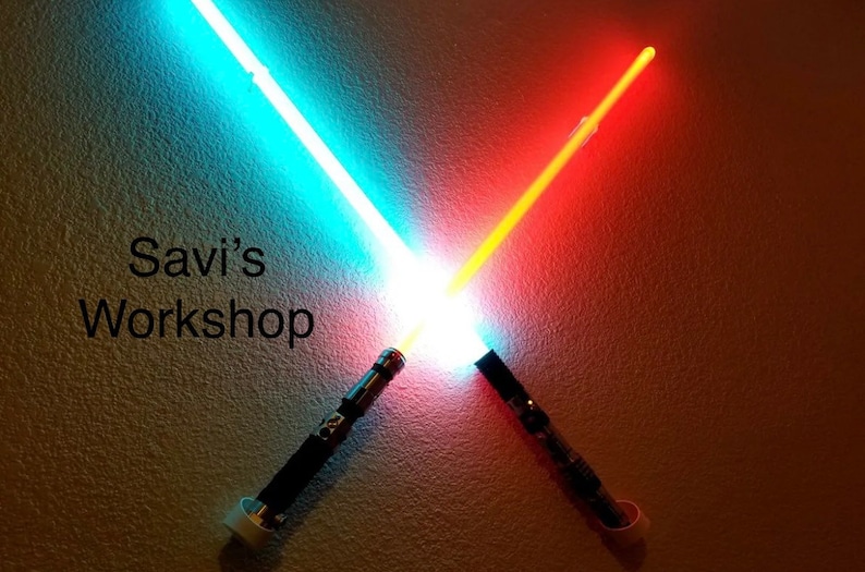May include: Two illuminated lightsabers, one blue and one red, are crossed against a textured wall. The text "Savi's Workshop" is displayed on the wall. The lightsabers have black handles.