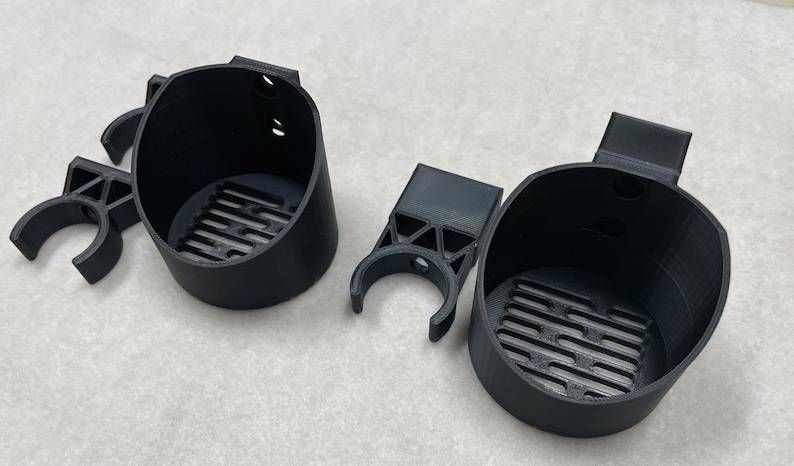May include: Black 3D-printed cup holders with a slotted base and a clip-on design. The holders have a cylindrical shape with a small handle on the side. The set includes two cup holders and two clip-on attachments.