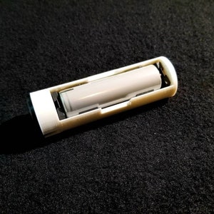 May include: A white rectangular battery holder with a gray cylindrical battery inside. The holder has a slot for the battery and is made of plastic. The background is a dark, textured surface.