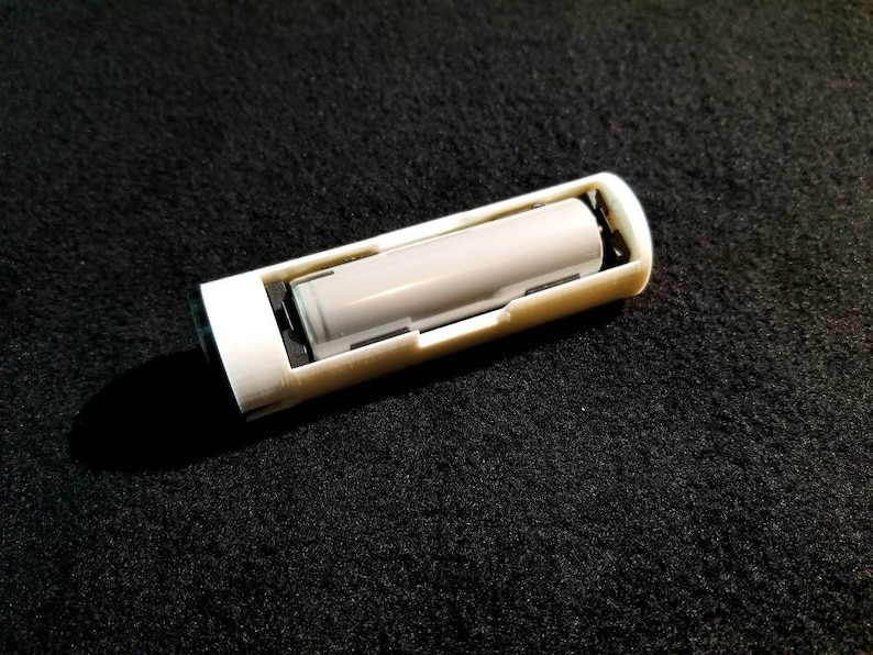 May include: A white rectangular battery holder with a gray cylindrical battery inside. The holder has a slot for the battery and is made of plastic. The background is a dark, textured surface.