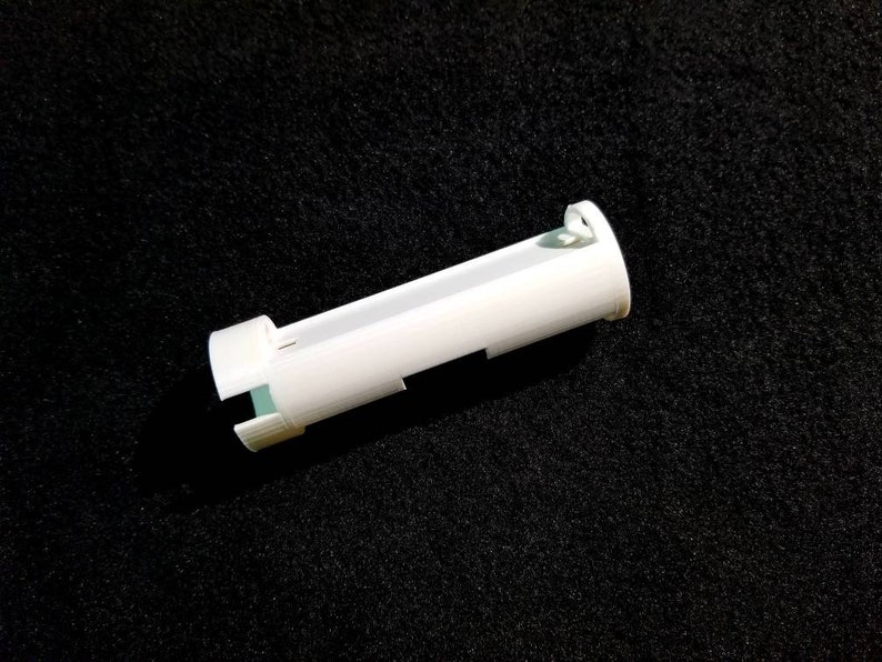 May include: A white, cylindrical plastic object with a rectangular opening and a small, rounded end. The object has a series of small, rectangular cutouts along its length. The object is lying on a black, textured surface.