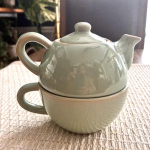 May include: A sage green ceramic tea-for-one set. The set includes a small teapot with a lid and a matching teacup. The teapot sits atop the teacup, creating a compact and elegant design. The set is ideal for a single serving of tea.
