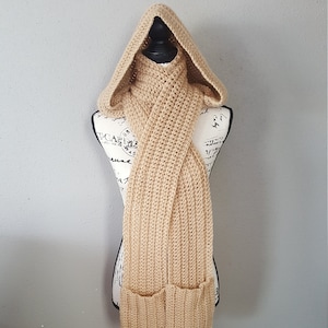 May include: A tan crocheted scarf with a hood and two pockets. The scarf is displayed on a black mannequin.