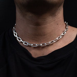 Puede incluir: Un collar de cadena de plata con un diseño de cadena de caja. La cadena la lleva una persona con una camisa negra.