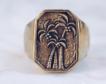 May include: A gold signet ring with an octagonal face. The face features a detailed relief of three palm trees. The ring has a polished finish and a solid appearance, suggesting a vintage or handcrafted style. The ring is likely made of gold.