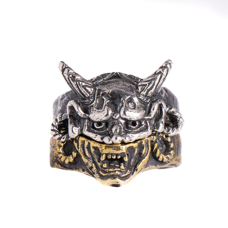 Puede incluir: Un anillo de plata y oro con un dise&ntilde;o detallado de una m&aacute;scara de demonio japon&eacute;s. La m&aacute;scara tiene cuernos, una expresi&oacute;n feroz y detalles intrincados.