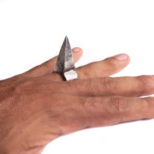 May include: A silver ring with a pointed, triangular design. The ring is worn on a person's finger.