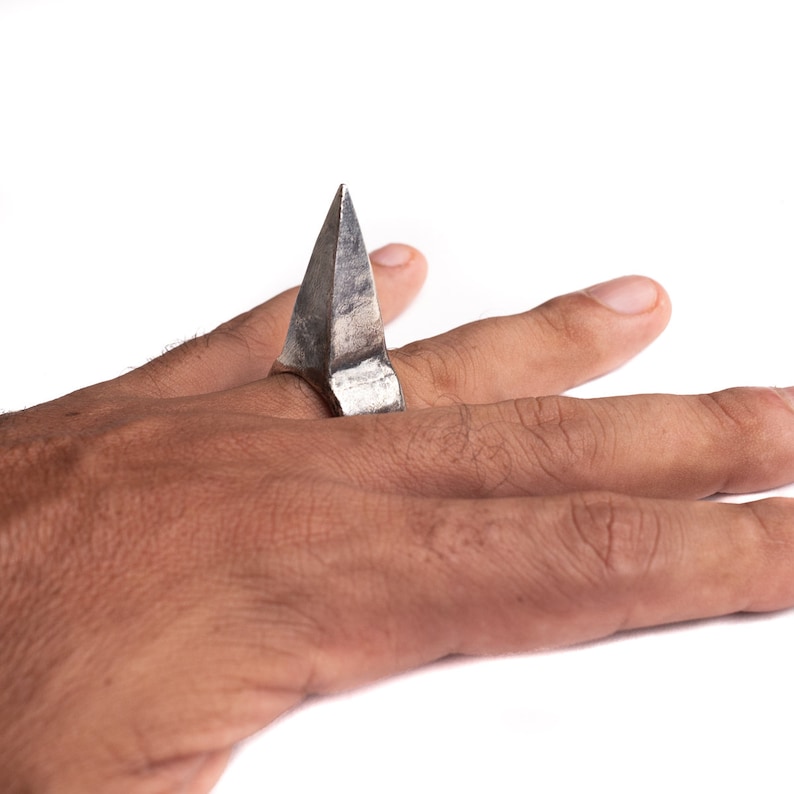 May include: A silver ring with a pointed, triangular design. The ring is worn on a person's finger.