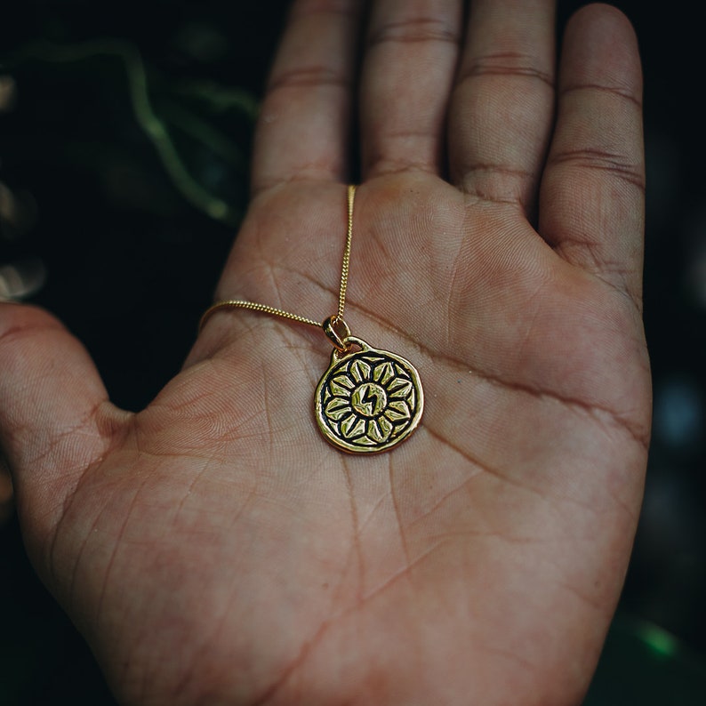 May include: A gold necklace with a round pendant featuring a stylized flower design with a lightning bolt in the center.