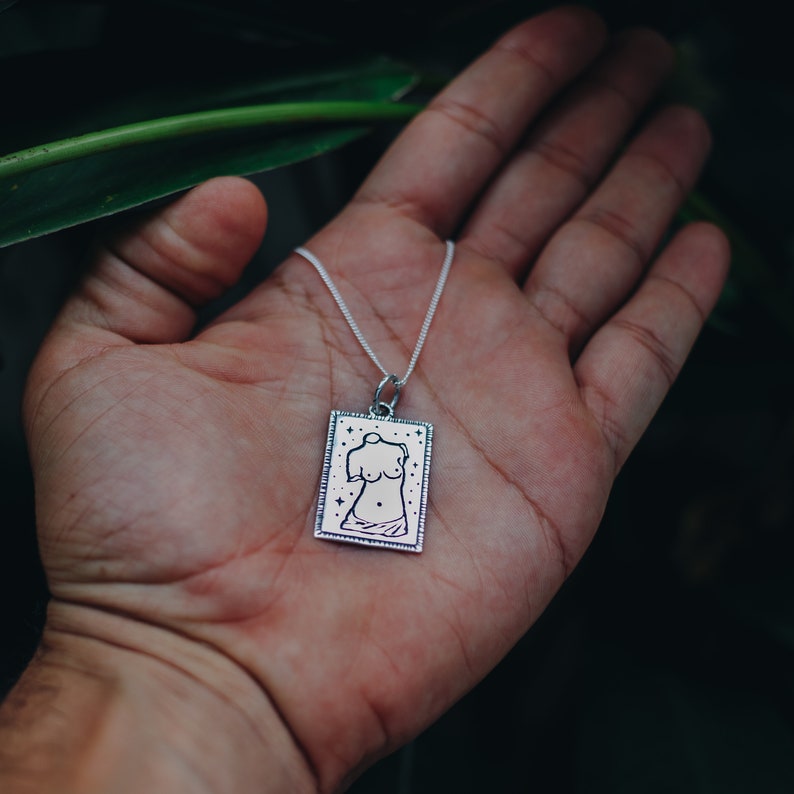 May include: A silver necklace with a rectangular pendant featuring a line drawing of a female torso with stars. The pendant has a textured border.