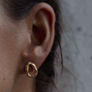 Peut inclure: Boucles d'oreilles cr&eacute;oles en or avec une finition textur&eacute;e martel&eacute;e. Les boucles d'oreilles sont petites et d&eacute;licates, et elles sont montr&eacute;es port&eacute;es &agrave; l'oreille d'une personne.