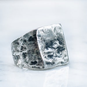 Könnte beinhalten: Ein silberner Ring mit einem strukturierten, rechteckigen Design. Der Ring hat eine raue, gehämmerte Oberfläche.