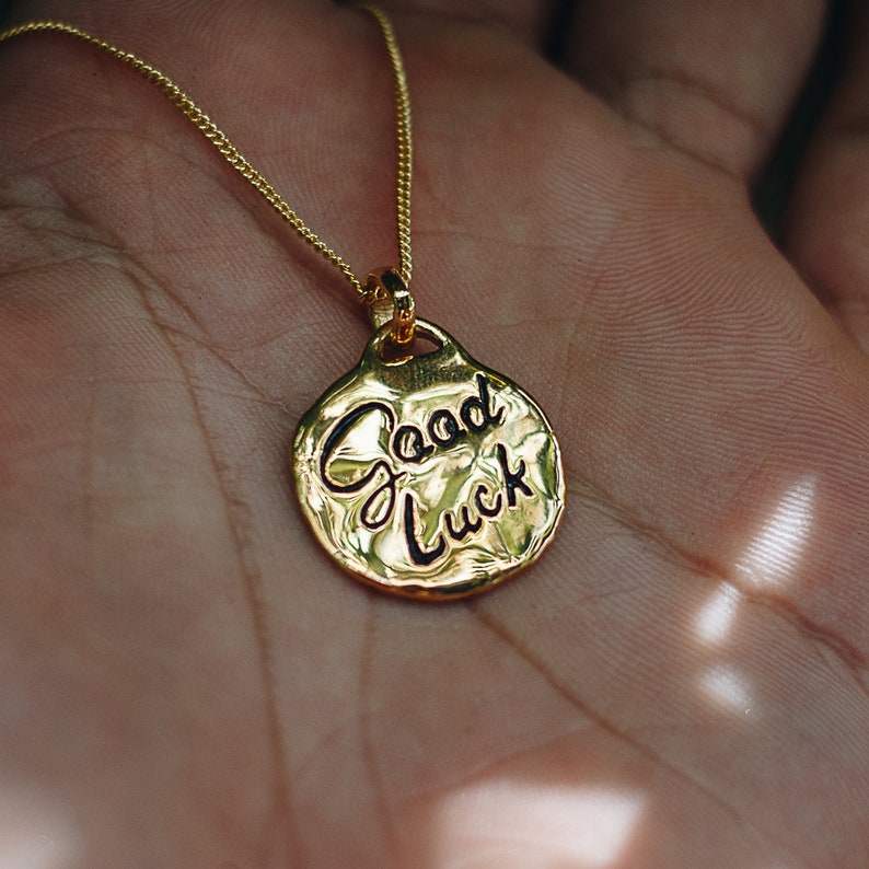May include: A gold necklace with a round pendant that says "Good Luck" in black lettering.