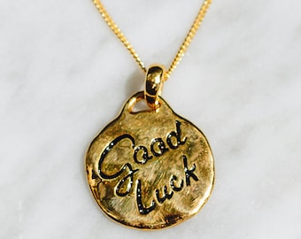 May include: A gold necklace with a round pendant that says "Good Luck" in black lettering.